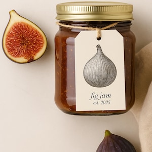 Op de afbeelding: Een pot vijgenjam met een gouden deksel en een label met een vijgenillustratie. Op het label staat "fig jam est. 2025". Een gehalveerde vijg en een hele vijg zijn ook zichtbaar.
