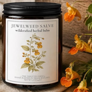 May include: A glass jar of Jewelweed Salve with a black lid, labeled "wildcrafted herbal balm." The label features a botanical illustration. The jar sits on a linen cloth, with orange flowers and a wooden bowl in the background. Net wt. 5 oz.