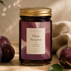 Op de afbeelding: Een glazen pot pruimenjam met een gouden metalen deksel en een etiket met de tekst The Thrifted Farm, Plum Preserve, small batch made by hand, 9 oz | 255 g. Het etiket heeft een paars pruimenpatroon. Drie hele donkerpaarse pruimen liggen ernaast.