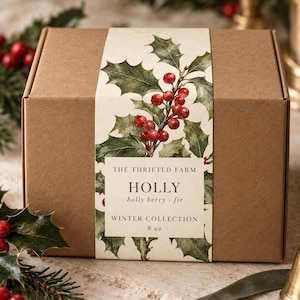 Op de afbeelding: Een rechthoekige bruine kartonnen doos met een papieren wikkel voorzien van een aquarelillustratie van groene hulstbladeren en rode bessen. De tekst luidt THE THRIFTED FARM HOLLY holly berry fir WINTER COLLECTION 9 oz in zwarte letters.
