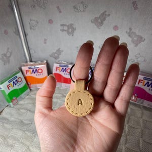 May include: A tan leather keychain with the letter "A" embossed in the center. The keychain has a stitched border and is attached to a black ring. Packages of FIMO soft polymer clay in various colors are in the background.