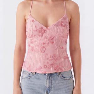 May include: A light pink camisole top with a V-neck and spaghetti straps. The top is embellished with floral embroidery and beadwork. The model is wearing light blue jeans.