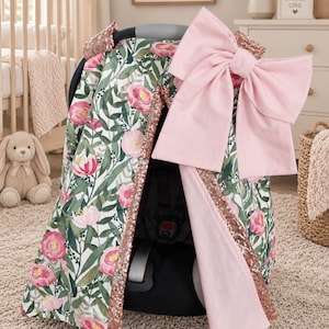 Può includere: Una copertura per seggiolino auto con un motivo floreale di peonie rosa e foglie verdi su sfondo bianco. La copertura è rifinita con bordi di paillettes color oro rosa e include un grande fiocco in tessuto rosa chiaro fissato sulla parte superiore.