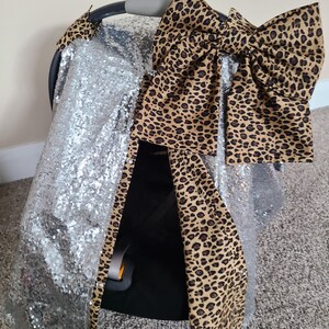 May include: A clear plastic car seat cover with a large brown and black leopard print bow on top. The cover is decorated with silver glitter.