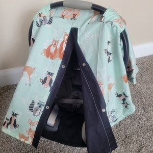 May include: A light blue and black car seat cover with a woodland animal print featuring deer, owls, raccoons, and bears. The cover has a black flap that can be used to cover the baby.