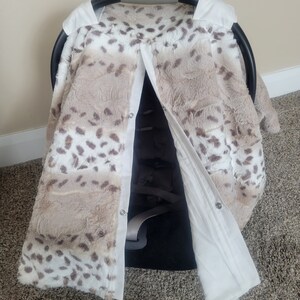 May include: A white and brown faux fur car seat cover with a leopard print pattern. The cover has a white lining and snaps to secure it to the car seat.