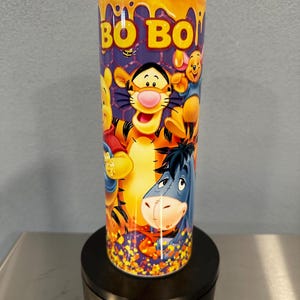 May include: A cylindrical tumbler with a clear lid featuring a colorful graphic print of cartoon characters including a tiger, a donkey, and a bear. The design includes yellow honey drips at the top, a purple honeycomb background, and the text BO BO.