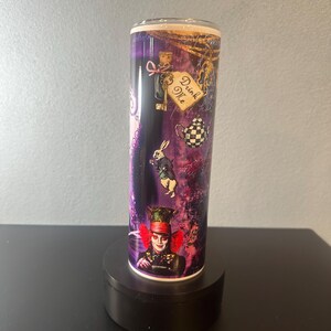May include: A cylindrical tumbler featuring illustrations from Alice in Wonderland. The design includes the Mad Hatter, the White Rabbit, and a "Drink Me" label, set against a purple and gold background. The tumbler is displayed on a black stand.