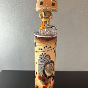 May include: A tumbler featuring a Funko Pop figure of a character from the board game Clue. The tumbler has a design with a fingerprint, magnifying glass, and the text "THE CLUE". The figure is standing on a clear lid.