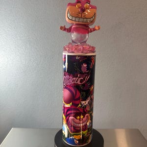 May include: A tumbler featuring a colorful design with the Cheshire Cat from Alice in Wonderland. The tumbler is topped with a pink Cheshire Cat figurine. The word "Magic" is written on the tumbler.