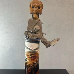 May include: A Star Wars-themed tumbler featuring Chewbacca and an AT-ST Walker. The tumbler has a brown and black design with a Chewbacca Funko Pop figure and AT-ST Walker toy attached to the top. The tumbler is cylindrical and sits on a black base.