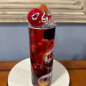 May include: A red and black tumbler featuring a football theme. The tumbler has a red helmet and a football on top. The design includes the team's logo and a red and black cloud-like pattern.