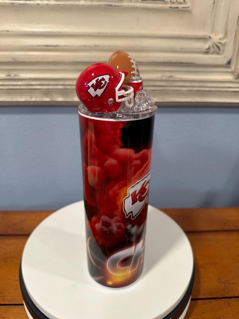May include: A red and black tumbler featuring a football theme. The tumbler has a red helmet and a football on top. The design includes the team's logo and a red and black cloud-like pattern.