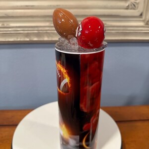 May include: A tall tumbler with a football theme. The tumbler features a brown football and a red helmet on top. The body of the tumbler has a fiery design with a football graphic. The tumbler is set on a white surface.