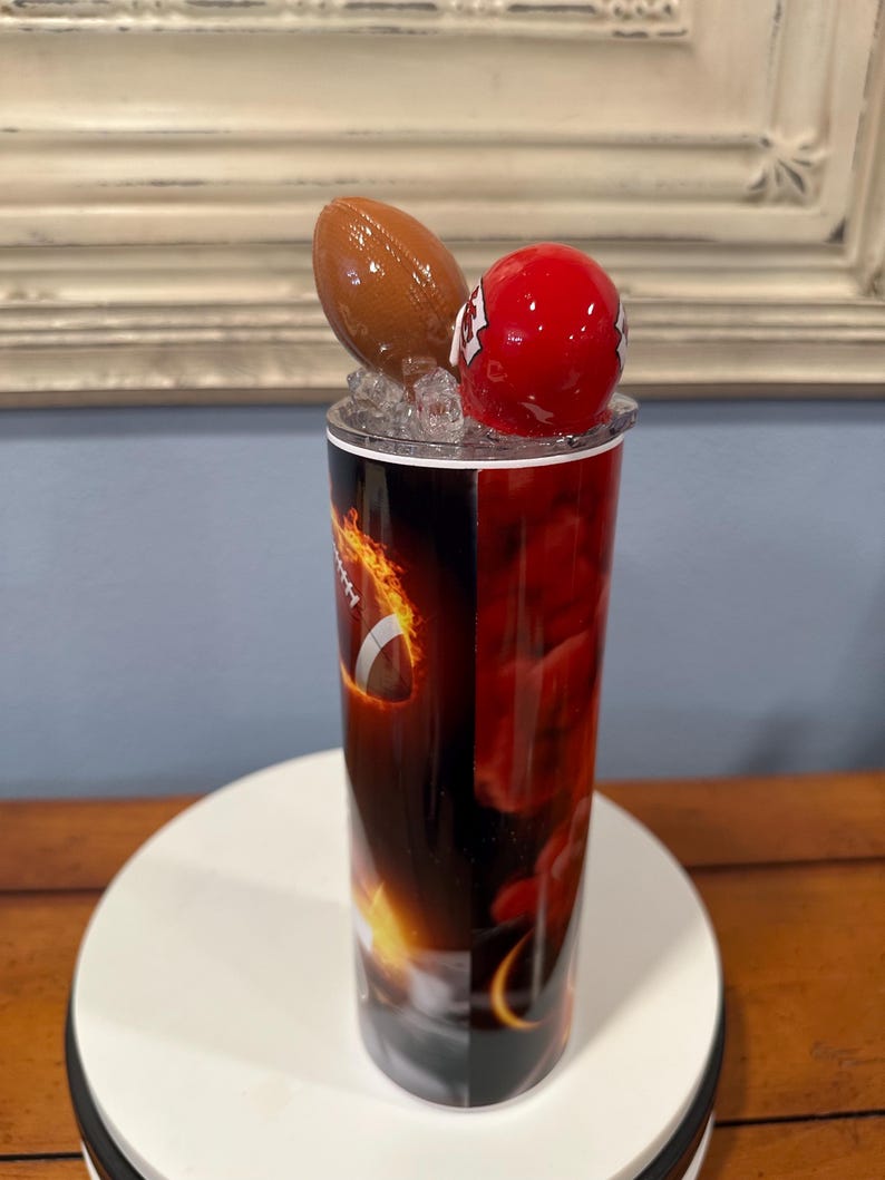 May include: A tall tumbler with a football theme. The tumbler features a brown football and a red helmet on top. The body of the tumbler has a fiery design with a football graphic. The tumbler is set on a white surface.