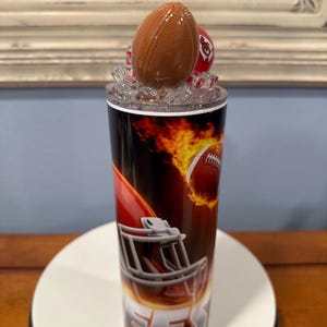 May include: A stainless steel tumbler with a football theme. The design features a football, a helmet, and flames. The lid is topped with a football-shaped bottle stopper and a red helmet. The tumbler is cylindrical and has a black, red, and orange color scheme.
