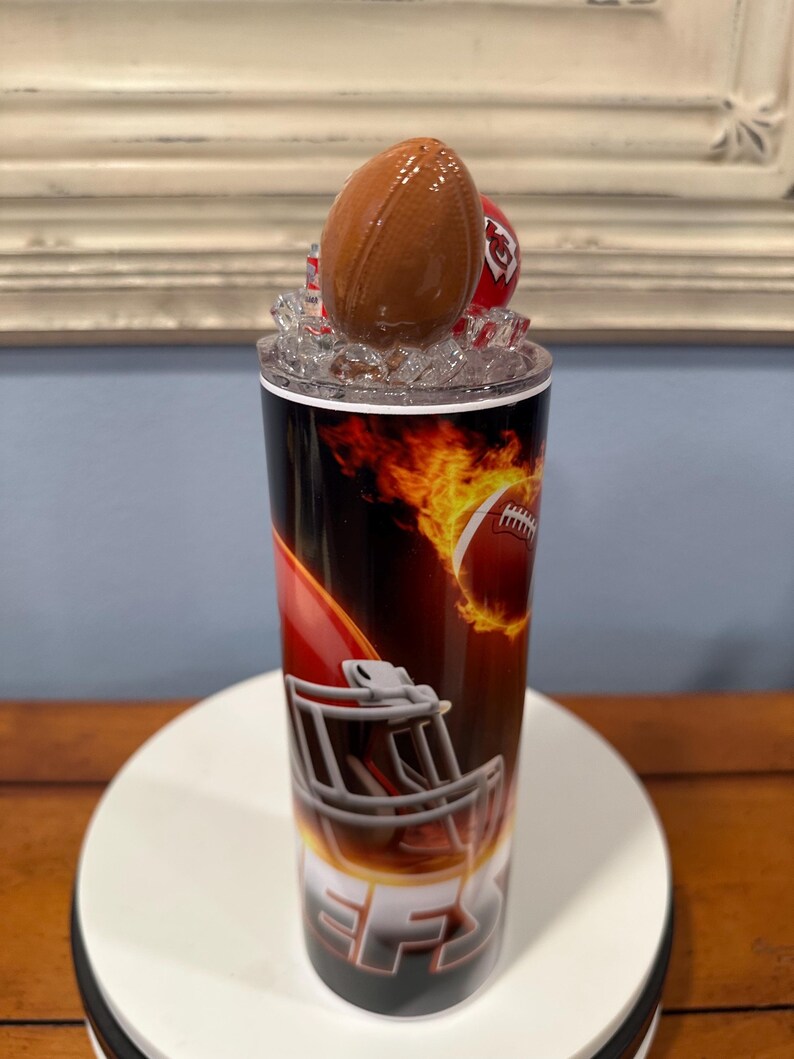 May include: A stainless steel tumbler with a football theme. The design features a football, a helmet, and flames. The lid is topped with a football-shaped bottle stopper and a red helmet. The tumbler is cylindrical and has a black, red, and orange color scheme.