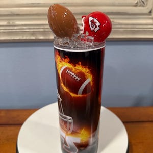 May include: A football-themed tumbler with a brown football and a red helmet on top. The tumbler features a football design with flames. The design is printed on a black background. The tumbler is on a white surface.