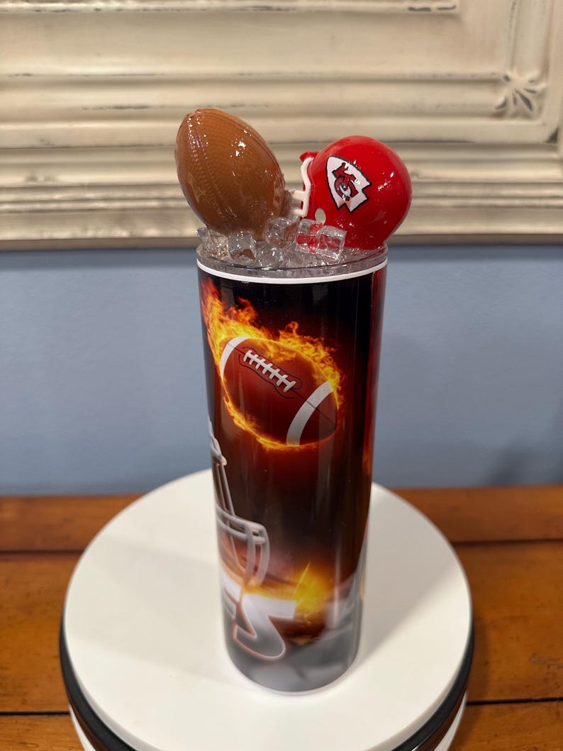 May include: A football-themed tumbler with a brown football and a red helmet on top. The tumbler features a football design with flames. The design is printed on a black background. The tumbler is on a white surface.