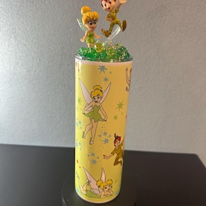 Peut inclure: Un gobelet jaune avec des illustrations de Clochette et Peter Pan. Le gobelet est surmonté de figurines de Clochette et Peter Pan. Le design comprend des étoiles et d'autres personnages du film. Le gobelet est sur une surface noire.