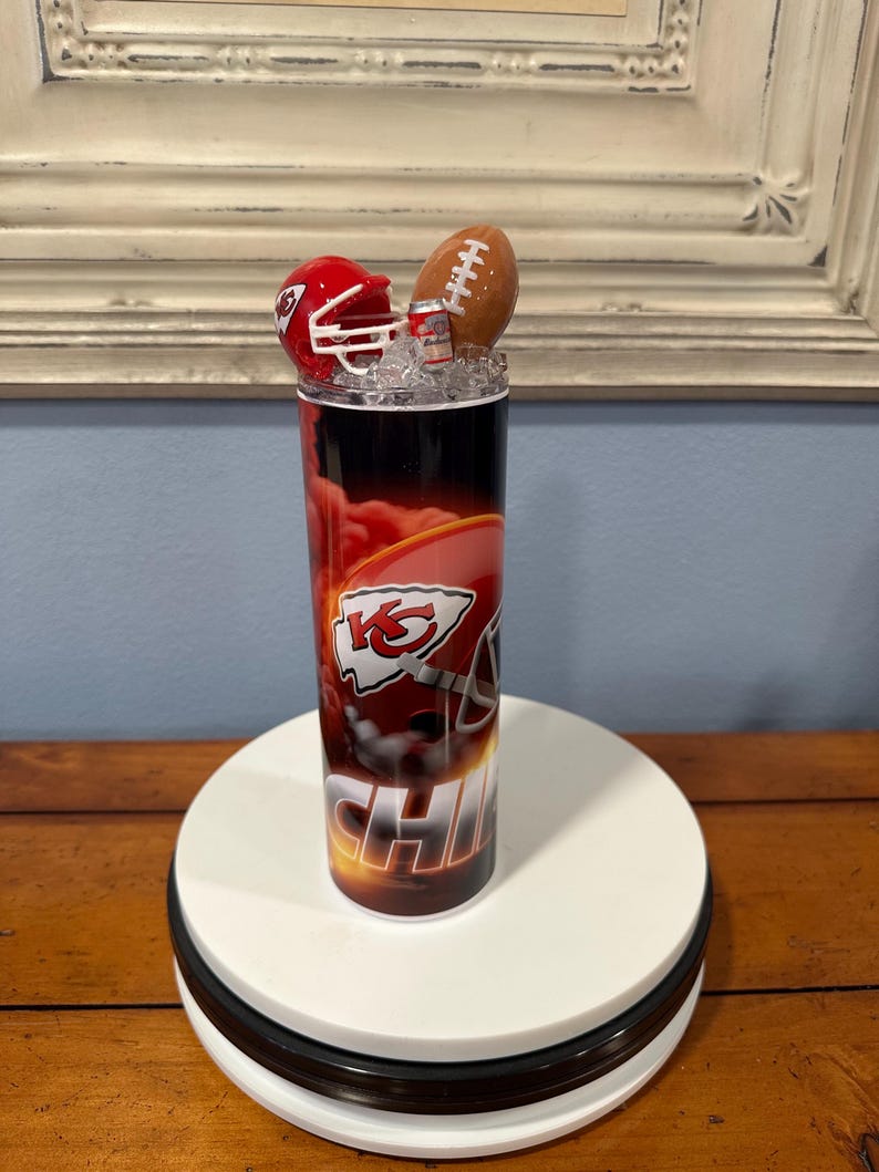 May include: A tall, cylindrical tumbler featuring a Kansas City Chiefs theme. The design includes the team's logo, a red helmet, and the word "CHIEFS". The top is decorated with a miniature football, a red helmet, and ice.