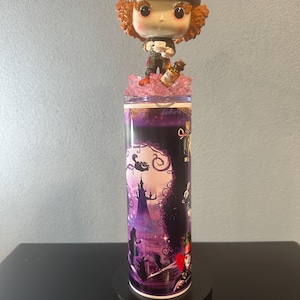 May include: A tall tumbler featuring a Mad Hatter Funko Pop figure on top. The tumbler has a purple and black design with images from Alice in Wonderland. The Mad Hatter figure wears a green hat with a pink ribbon.
