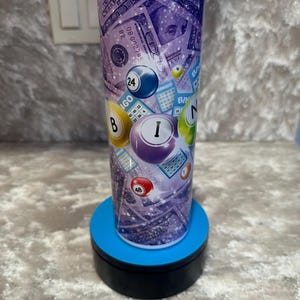 May include: A tall, cylindrical tumbler with a purple and white design featuring dollar bills and bingo balls. The tumbler has a clear lid and is set on a blue and black rotating stand. Bingo balls with numbers like 24, 48, and I are visible.