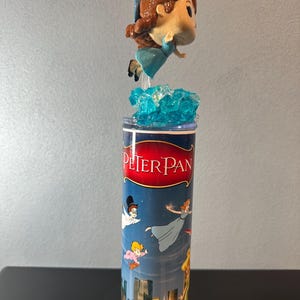 Peut inclure: Un gobelet sur le thème de Peter Pan avec une figurine d'un personnage en robe bleue et aux cheveux bruns. Le gobelet présente les mots "Peter Pan" et des illustrations de personnages volant au-dessus d'une ligne d'horizon urbaine. La figurine est suspendue au-dessus d'une base bleue texturée.