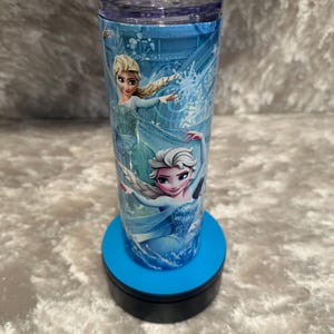 May include: A blue tumbler featuring images of Elsa from the animated film Frozen. The tumbler has a clear lid and is set on a blue and black base. The design includes Elsa in a flowing blue dress, with a wintery background.