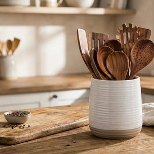 May include: A selection of wooden kitchen utensils, such as spoons and spatulas, are arranged in a white ceramic utensil holder. The kitchen scene includes a wooden chopping board, bowls, and a bottle of oil, creating a warm and inviting atmosphere.