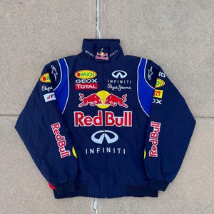 以下が含まれることがあります： Red Bull、Infiniti、Geox、Total、Pepe Jeansのロゴが入ったネイビーブルーのジップアップジャケット。フロントには大きな赤と黄色の雄牛のロゴが描かれ、袖と胸にはさまざまなスポンサーのワッペンやロゴが施されています。
