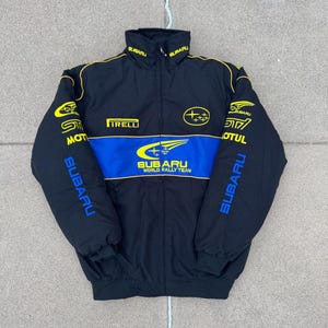 Könnte beinhalten: Eine schwarze Jacke mit Reißverschluss und einem blauen Querstreifen auf der Brust mit dem Text Subaru World Rally Team. Die Jacke zeigt gelbe Subaru-Logos, Pirelli- und Motul-Schriftzüge sowie das Wort Subaru auf beiden Ärmeln.