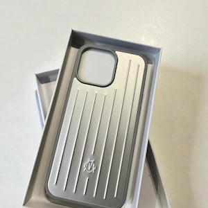 Peut inclure: Un étui de téléphone argenté et gris avec un design strié, présenté dans une boîte blanche. L'étui a une bordure grise et une découpe pour l'appareil photo. Un petit emblème est visible sur la partie inférieure de l'étui.
