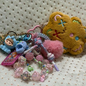 May include: A collection of keychains and accessories, including a plush gingerbread man, a pink pom-pom, and a bracelet with pink and clear beads. Other items include a blue baby figurine and a pink and purple keychain.