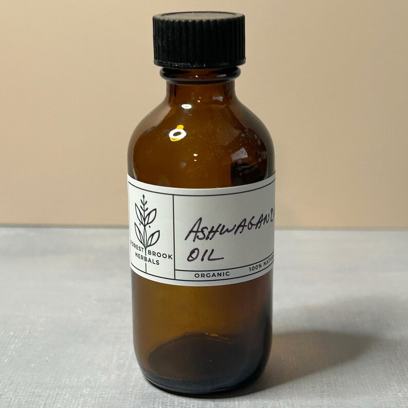 May include: A brown glass bottle with a black cap, labeled "Ashwagandha Oil" and "Organic." The label also features the text "Forest Brook Herbals" and a botanical illustration. The bottle is filled with a golden liquid.