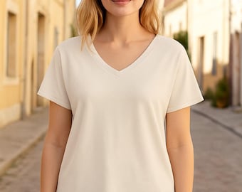 Könnte beinhalten: Ein cremefarbenes V-Ausschnitt-T-Shirt mit kurzen Ärmeln. Das Shirt wird mit einer blauen Jeans kombiniert. Das Model steht vor einem verschwommenen Hintergrund einer europäischen Straße.