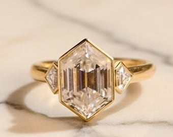 May include: A gold ring with a large, clear, hexagonal-cut center stone, flanked by two smaller, triangular-cut stones. The ring is set on a marble surface, showcasing the ring's design and sparkle.