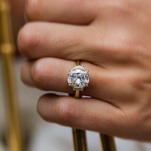 May include: A gold ring with a large, cushion-cut diamond. The ring is on a person's finger, with a white purse and gold handle in the background. The diamond is clear and sparkling.