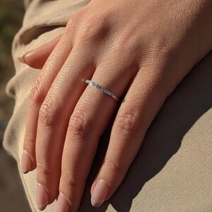 May include: A thin silver-toned metal band ring featuring a series of small, rectangular baguette-cut clear stones set horizontally along the top half of the band. The ring has a polished finish and a slim, delicate profile on the finger.