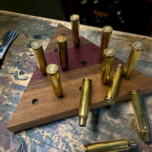 May include: A triangular wooden display stand featuring a multi-toned wood grain pattern with several upright brass bullet casings inserted into pre-drilled holes, with one additional brass casing lying horizontally on the surface.