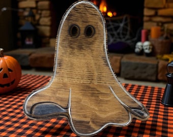 May include: Wooden ghost-shaped serving tray with a silver-colored trim. The tray has two round eye cutouts and a carved mouth. It is placed on a black and orange checkered tablecloth, with a carved pumpkin and fireplace in the background. A Halloween-themed decorative item.