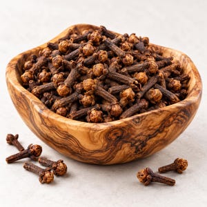 May include: A wooden bowl with a distinct swirling grain pattern filled with a large quantity of dried whole cloves. The cloves are dark brown, featuring a narrow stem and a rounded, textured flower bud at the top. Several loose cloves lie nearby.