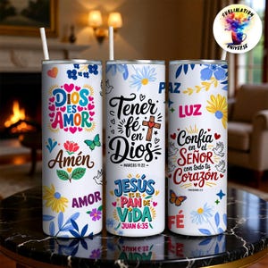 May include: Three white insulated tumblers with floral and butterfly designs. Each tumbler features religious text in Spanish, including "Dios es Amor" and "Jes&uacute;s es el Pan de Vida." The tumblers have a straw and are decorated with colorful flowers.