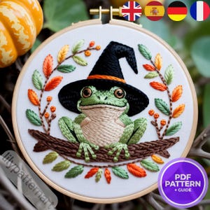 Peut inclure: Broderie d'une grenouille verte portant un chapeau de sorcière, assise sur une branche, entourée de feuilles d'automne. La grenouille a un ventre beige et un chapeau noir avec une bordure orange. Le texte "PDF PATTERN + GUIDE" est visible.