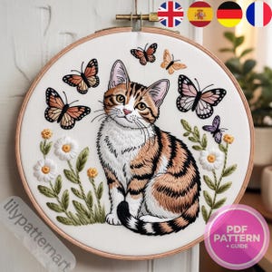 Puede incluir: Bordado detallado de un gato atigrado con mariposas y flores. El gato tiene rayas marrones y naranjas, con detalles blancos. Mariposas en tonos naranja, negro y morado rodean al gato. La obra de arte está en un bastidor de bordado de madera.