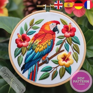 Puede incluir: Arte bordado en bastidor que presenta un loro vibrante con plumaje rojo, naranja, amarillo y azul, posado en una rama con flores de hibisco y hojas verdes. La obra de arte está sobre un fondo blanco dentro de un bastidor de madera.