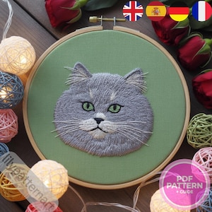 Puede incluir: Un rostro de gato bordado en un aro de madera. El gato es gris con ojos verdes y bigotes blancos, sobre una tela verde. El aro está rodeado de luces de cadena y rosas rojas. El texto "PDF PATTERN + GUIDE" está en un círculo rosa.
