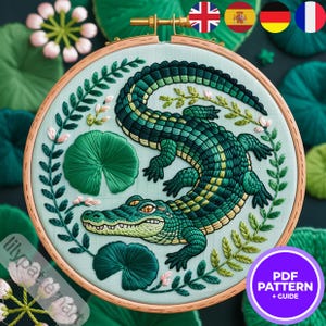 Peut inclure: Broderie sur tambour représentant un alligator vert détaillé entouré de feuilles vertes et de fleurs roses. Le fond est bleu clair. Le texte "PDF PATTERN + GUIDE" est dans un cercle violet, indiquant un patron et un guide.