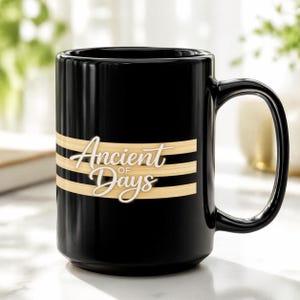 May include: Black ceramic mug with a large handle. The mug features the words "Ancient of Days" in white script over two horizontal tan stripes. The mug is on a white surface.