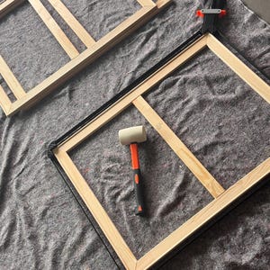 May include: Two rectangular light-colored wooden frames with a central support bar, a black nylon strap clamp secured around one frame, and a rubber mallet with an orange handle and white head resting in the center of the clamped frame.